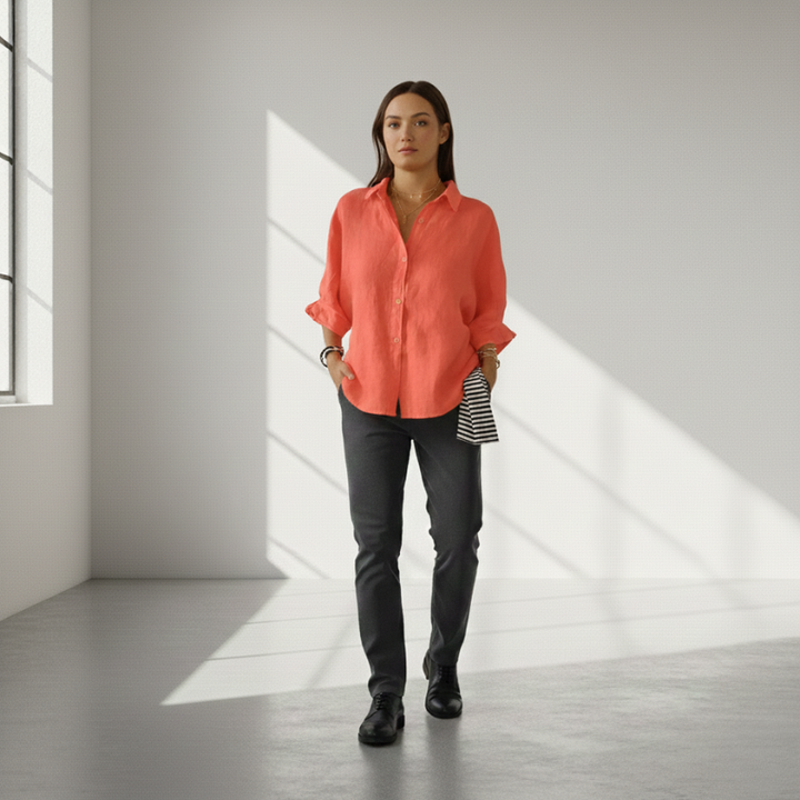 Woman wearing a coral shirt and dark pants standing in a well-lit room with sunlight streaming through a window.