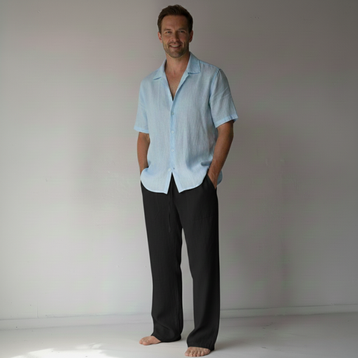 Man wearing a light blue shirt and black pants standing against a plain background