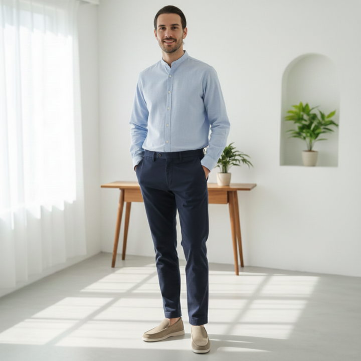 Man wearing a light blue shirt and dark pants standing in a bright room with a desk and plants.
