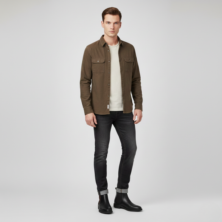 Man wearing a brown jacket, white shirt, black jeans, and black boots on a light gray background