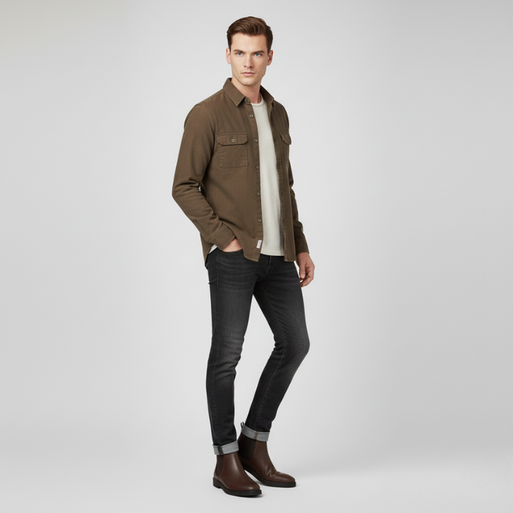 Man wearing a brown jacket, white shirt, black jeans, and brown boots on a light gray background