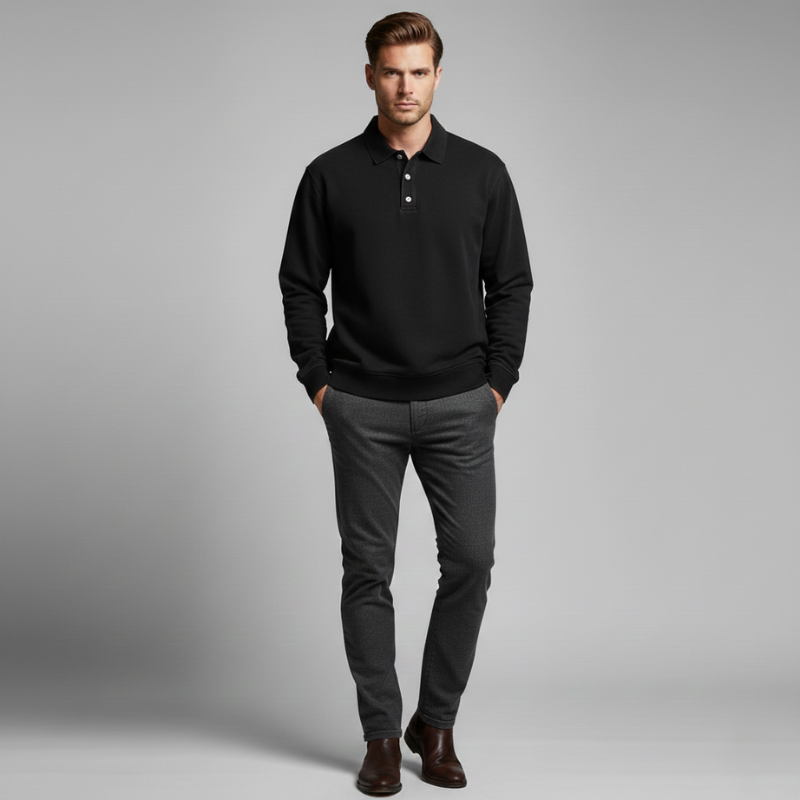 Man dressed in a black pullover and slim trousers standing in a plain light gray setting.