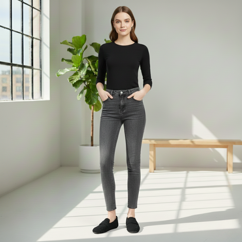 Woman wearing a black top and gray jeans standing in a bright room with a plant and bench.