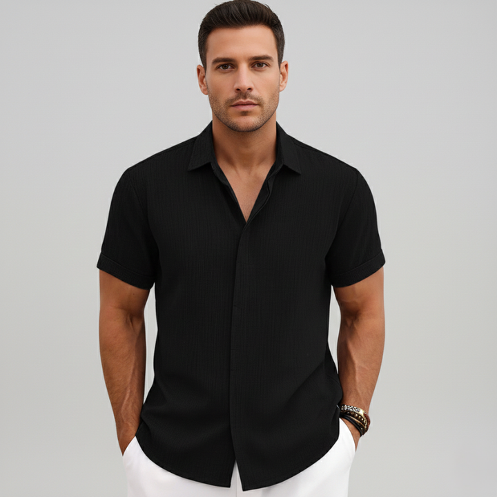 Man standing indoors wearing a black short-sleeve shirt paired with light gray trousers in natural lighting.