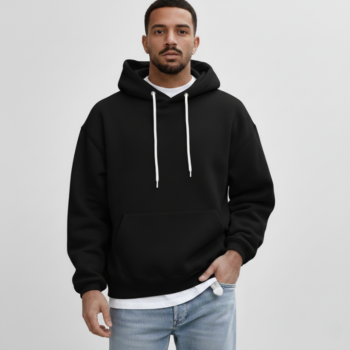 Man wearing a black fleece pullover hoodie with white drawstrings and a relaxed fit in a clean studio setting.