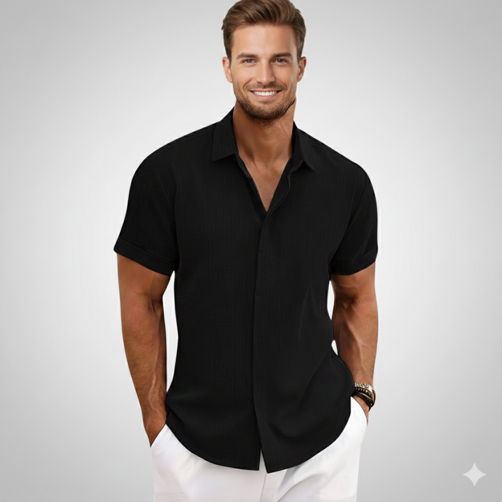 Man wearing a black casual short-sleeve shirt styled with white pants in soft natural indoor light.