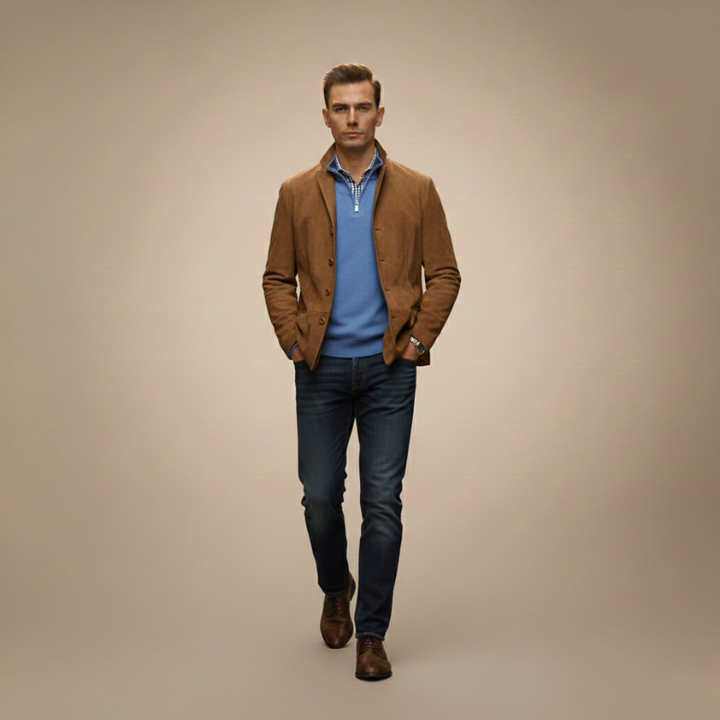 Man standing in warm studio lighting wearing a brown blazer jacket styled with jeans and dress shoes.