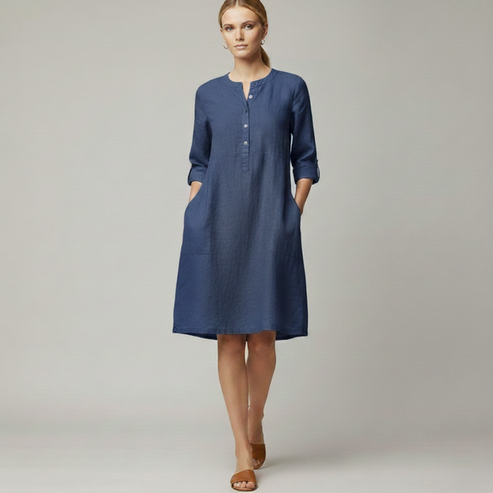 Female model standing indoors wearing a blue everyday dress with a simple, relaxed silhouette and soft fabric texture.