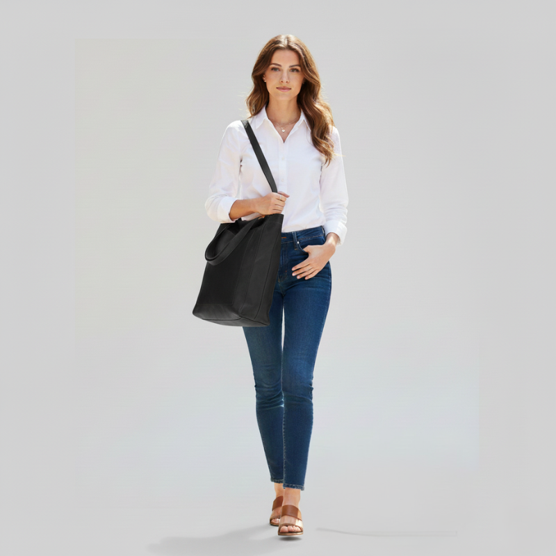 Woman standing with a brown leather tote bag in natural light, styled with a white blouse and denim jeans.