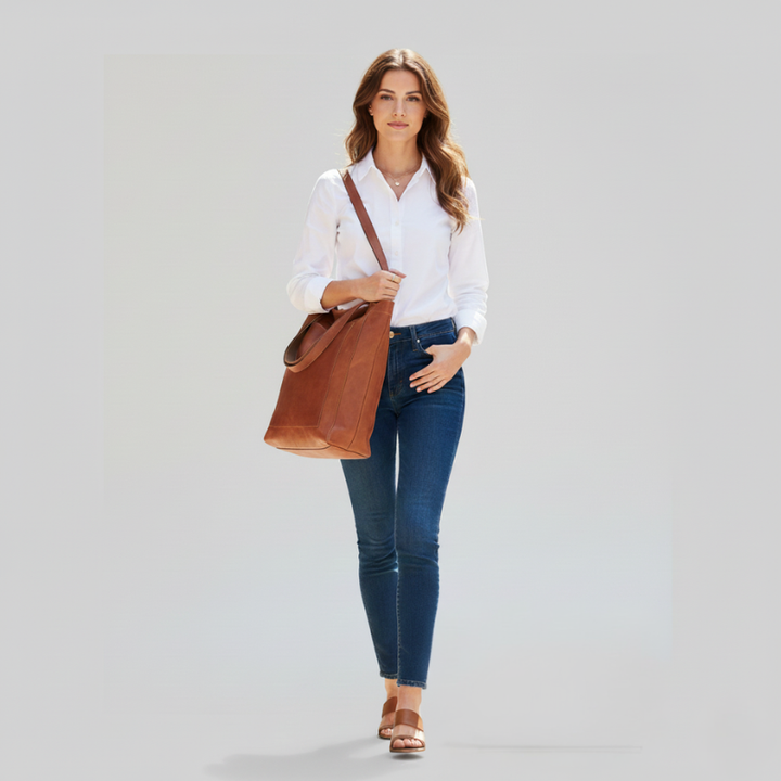 Woman carrying a brown leather tote bag paired with a white blouse and jeans in bright studio lighting.